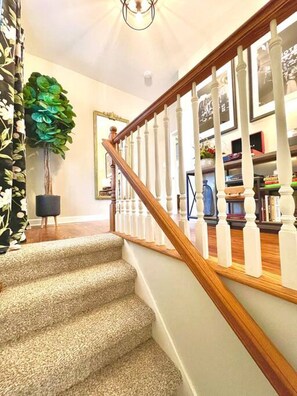 A carpeted staircase with a wooden handrail and white balusters, leading to a room with a bookshelf and a potted plant.