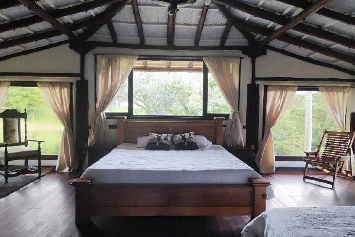 A bedroom with a wooden bed, two windows with curtains, a chair, and a view of greenery outside.