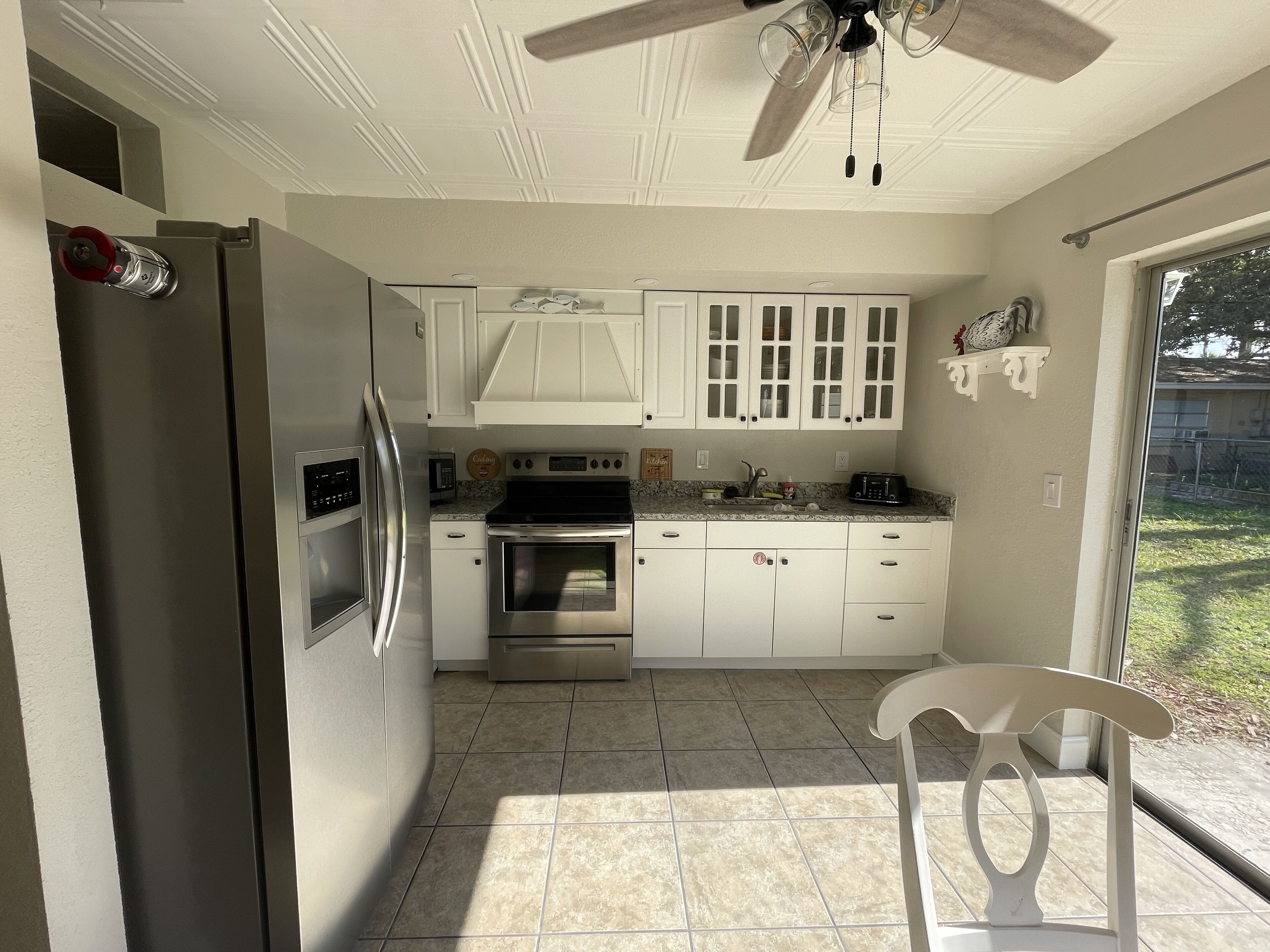 A compact kitchen with white cabinets, a stainless steel refrigerator, an oven, and a microwave. There is a ceiling fan, a window with a view of the outdoors, and a white chair in the corner.