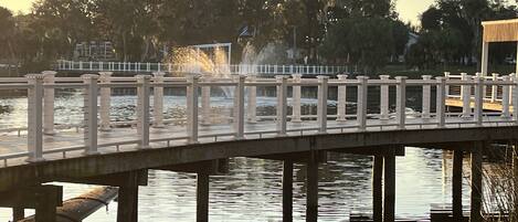 A bridge over water with a railing, a grassy area in the foreground, and trees in the background.