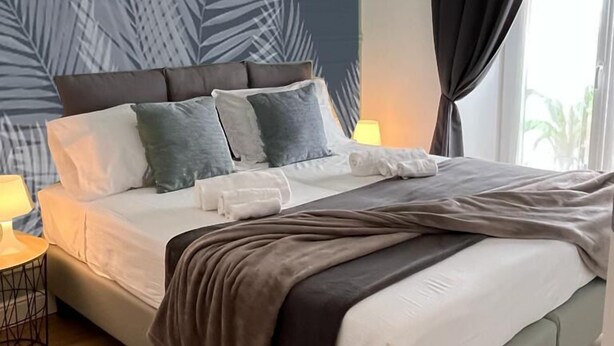Un lit bien fait, avec une tête de lit grise, des oreillers blancs et gris, et une couverture marron. Une lampe de chevet se trouve sur le côté gauche du lit.