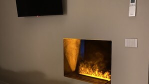 Et værelse med et fladskærms-tv monteret på væggen, en pejs med synlige flammer, et termostat og en stikkontakt.