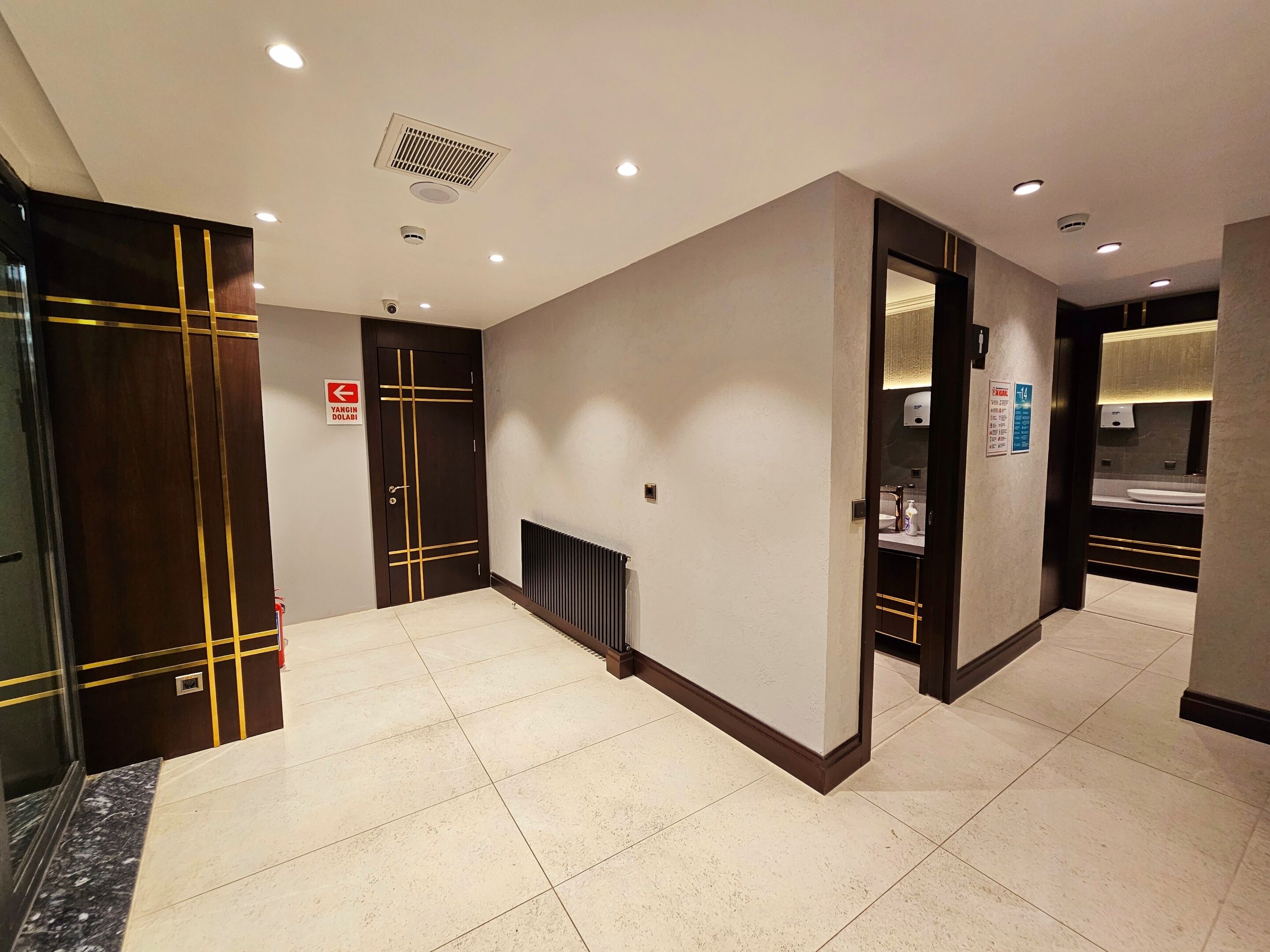 Vestíbulo de hotel con diseño moderno, que presenta una puerta de madera oscura con detalles dorados, suelo de baldosas y un radiador.