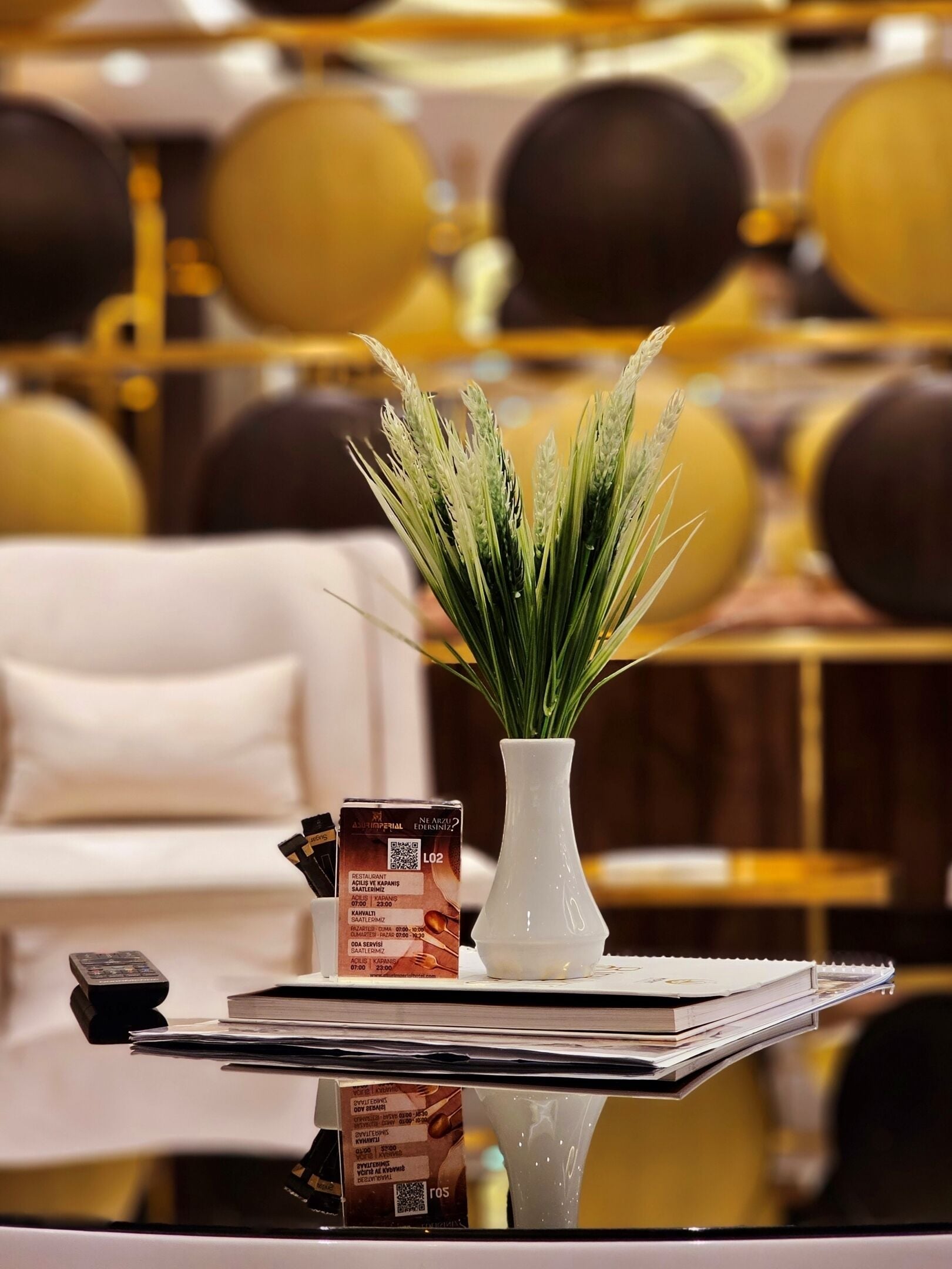 Une table sur laquelle se trouve un vase blanc contenant des plantes vertes, un menu et une télécommande.