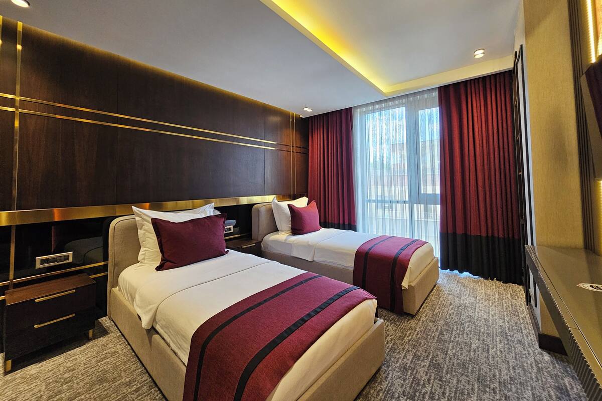 Une chambre d’hôtel avec deux lits, une tête de lit en bois foncé, une grande fenêtre avec des rideaux rouges et un sol recouvert de moquette.