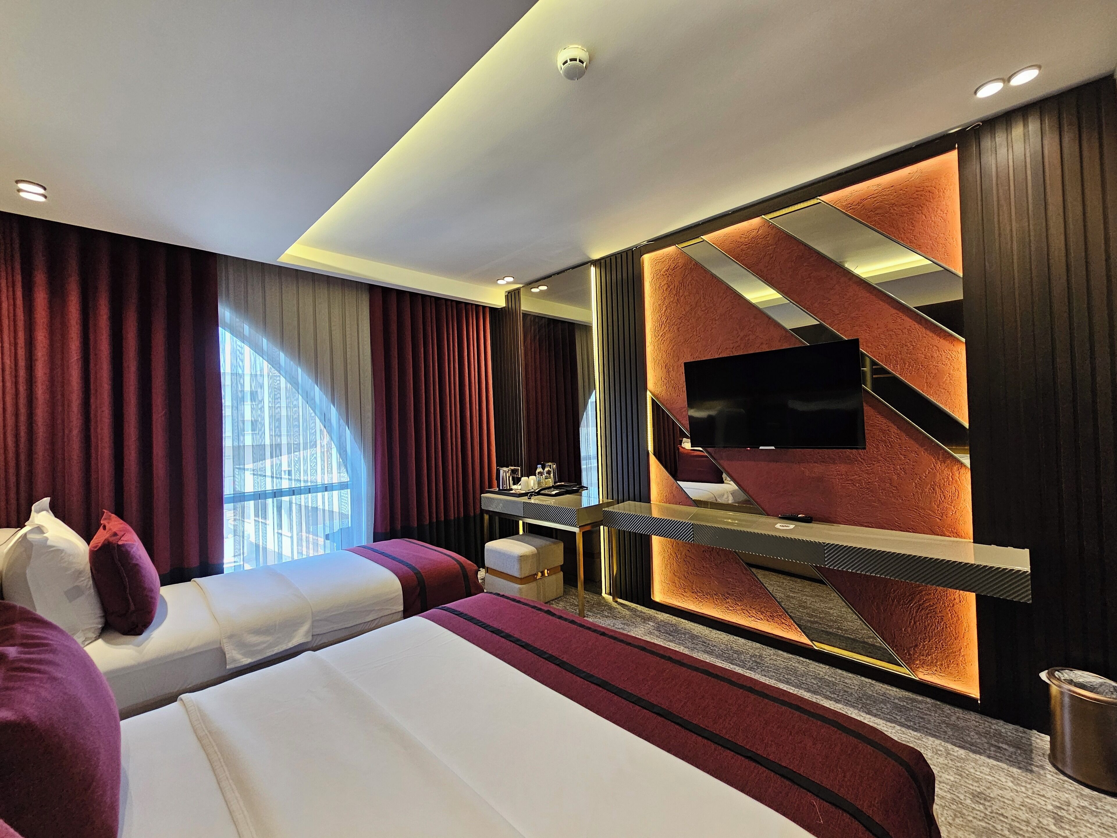 Una habitación de hotel moderna con una cama grande, un escritorio con televisión de pantalla plana y un amplio ventanal con cortinas.