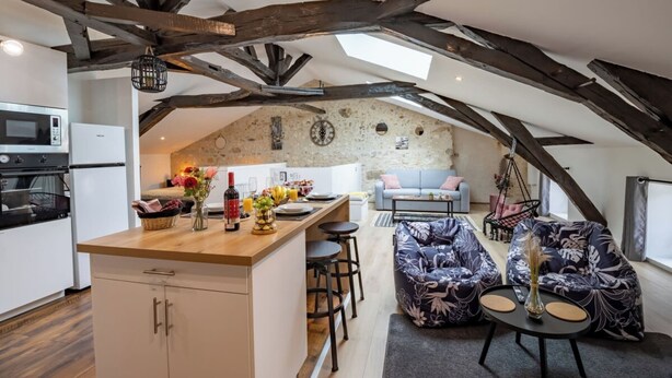 A cozy loft-style apartment with exposed wooden beams, a kitchen with modern appliances, a dining area with a hanging chair, and a living room with a sofa and a coffee table. - Le Loft - Rated 5 Stars (Mussidan)