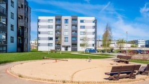A modern apartment building with a landscaped courtyard featuring a sandpit, wooden benches, and a seesaw. - HARAKA APARTMENT WITH SUNSET BALCONY  (Tartu)