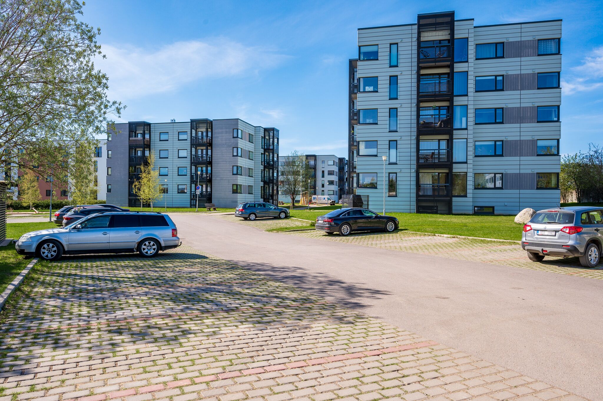 Un immeuble d’appartements moderne composé de plusieurs bâtiments, avec une allée pavée et des voitures garées.