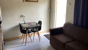 Una sala pequeña con un sofá marrón, una mesa de comedor con sillas, un cuadro enmarcado en la pared y una puerta con cortina.