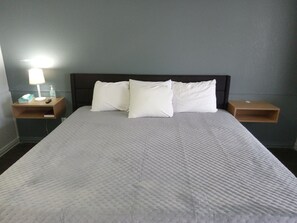 A neatly made bed with a white quilt, two white pillows, and a wooden headboard. There are two bedside tables, one with a lamp and a tissue box, and the other with a bottle of hand sanitizer. - Western Hills Motel (Versailles)