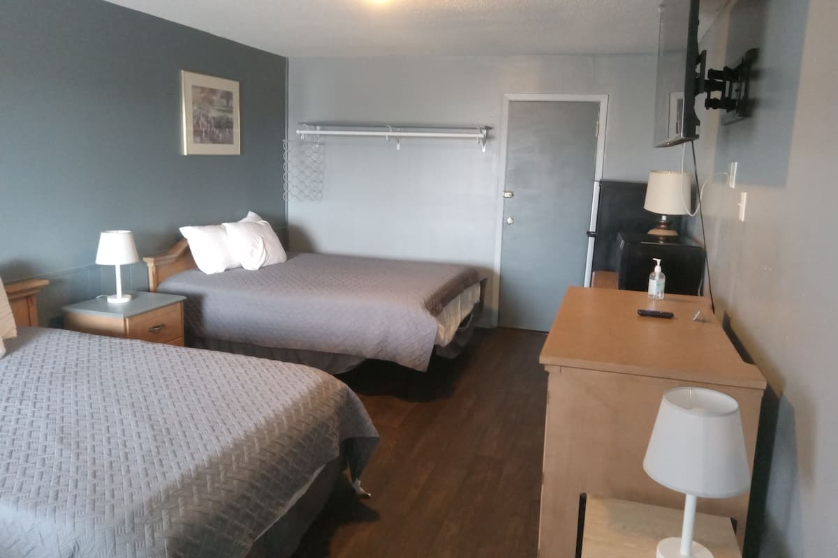 A hotel room with two beds, a wooden desk, a chair, a nightstand, a lamp, a door, and a wall-mounted TV.