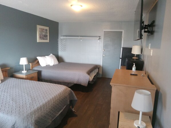 A hotel room with two beds, a wooden desk, a chair, a nightstand, a lamp, a door, and a wall-mounted TV. - Western Hills Motel (Versailles)