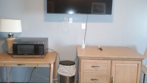 A small room with a television mounted on the wall, a microwave on a wooden table, and a small cabinet with drawers. - Western Hills Motel (Versailles)