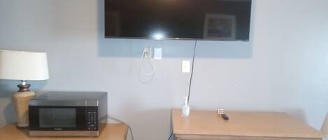 A small room with a television mounted on the wall, a microwave on a wooden table, and a small cabinet with drawers.