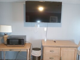 Una habitación pequeña con un televisor montado en la pared, un microondas sobre una mesa de madera y un mueble bajo con cajones.