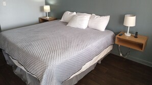 A neatly made bed with a grey quilt, two bedside tables with lamps, and a wooden nightstand with a remote control. - Western Hills Motel (Versailles)