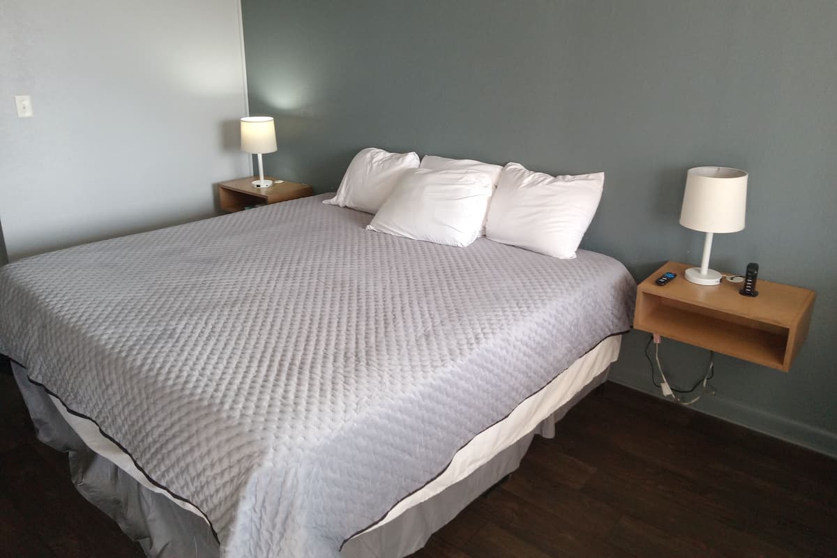 A neatly made bed with a grey quilt, two bedside tables with lamps, and a wooden nightstand with a remote control.
