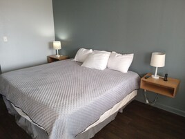 Una cama bien tendida con un colcha gris, dos mesitas de noche con lámparas y un mesero de madera con un control remoto.