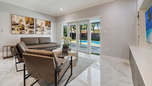 A modern living room with a sofa, armchairs, and a coffee table. There are abstract paintings on the wall and a view of a swimming pool through glass doors. - Tropical Miami 5BR Retreat • Game Room • Pool_BBQ (Miami)