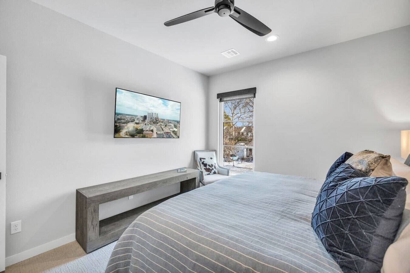 A bedroom with a bed, a ceiling fan, a mounted TV, a window with a view, and a bedside table.