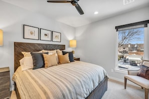 A bedroom with a bed, a ceiling fan, framed pictures on the wall, and a window with a view of the outside.