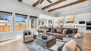 A spacious living room with a large brown leather sofa, a coffee table, and a dining area with a chandelier. - Lux 5BD Stonefront Tulsa | Near Gathering Place (Tulsa)