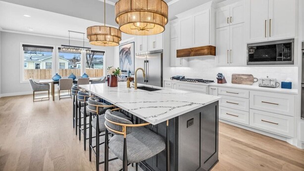 A modern kitchen with white cabinetry, a large island with bar stools, pendant lighting, and a view of the backyard. - Lux 5BD Stonefront Tulsa | Near Gathering Place (Tulsa)