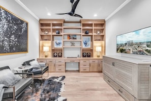 A modern living room with a built-in entertainment unit, a large flat-screen TV, a ceiling fan, and a large abstract painting.