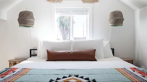 A bedroom with a bed, white bedding, a colorful blanket, two hanging lamps, and a window with curtains.