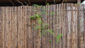 A wooden lounge chair with a slatted design, positioned on a concrete surface with a backdrop of a bamboo fence and a stone planter with rocks.