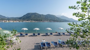 Ein Strand mit Reihen blauer Liegestühle und weißen Sonnenschirmen, ein Kiesstrand und ein Blick auf einen See mit Bergen im Hintergrund.