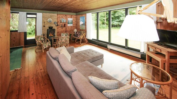 Een gezellige woonkamer met een grote bank, rieten stoelen, een open haard en grote ramen met uitzicht op de natuur.