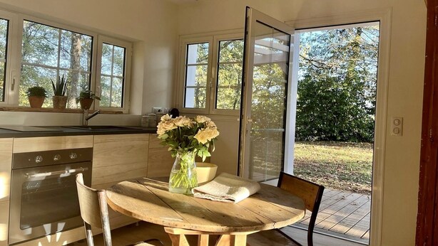Una cocina con una mesa y sillas de madera, un jarrón de flores y un patio al que se accede por un toldo de vidrio corredizo.