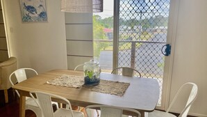 Ein Essbereich mit Holztisch, weißen Stühlen, einem Glasgefäß mit Pflanzen, einer Spitzen Tischmatte und Blick durch eine Drahtgittertür.