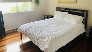 Ein ordentlich gemachtes Bett mit weißer, strukturierter Decke, einem dunklen Holz-Kopfteil, einem kleinen Nachttisch, einer hängenden Pflanze und einem Palmenbaumgemälde an der Wand.