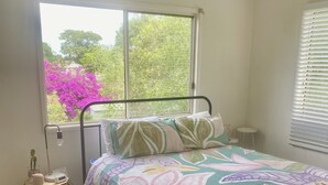 Ein Schlafzimmer mit Bett, Fensterblick auf Bäume und Blumen, Nachttisch mit Lampe sowie Deckenventilator.