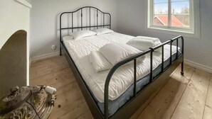 Un dormitorio con estructura de cama de metal, ropa de cama blanca, suelo de madera y ventana con vista a edificios.