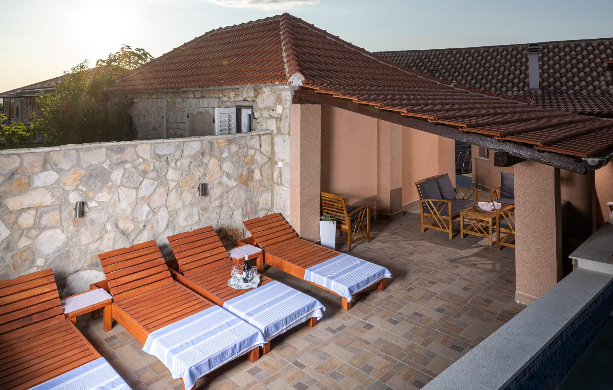 A rooftop terrace with wooden lounge chairs and tables, stone walls, and a tiled roof.
