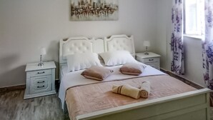Een net opgemaakt bed met kussens en een opgerolde handdoek, een nachtkastje met een lamp, en een schilderij aan de muur.