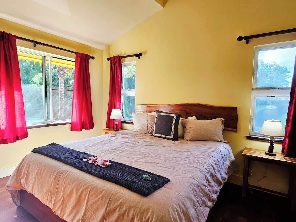 A neatly made bed with a headboard, two bedside lamps, and a window with red curtains.