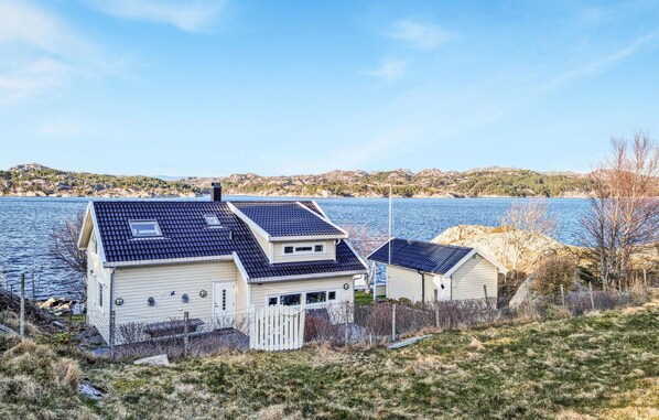 Il s’agit d’une propriété située en bord de mer, comprenant deux bâtiments principaux : un plus grand, doté d’un toit en pente et de panneaux solaires, et un plus petit, avec un toit plat. Les deux bâtiments sont pourvus de fenêtres bien visibles et sont entourés d’une cour clôturée.