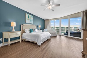 Une chambre à coucher comprenant un lit, des tables de chevet, une commode, un ventilateur de plafond et de grandes fenêtres donnant sur un immeuble.