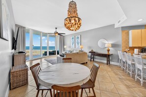 A spacious living area with a large round table, a dining area with white chairs, a kitchen with wooden cabinets, and a balcony with a view of the ocean. - Professionally Decorated Penthouse Unit! ASK ABOUT DISCOUNTS! (Orange Beach)