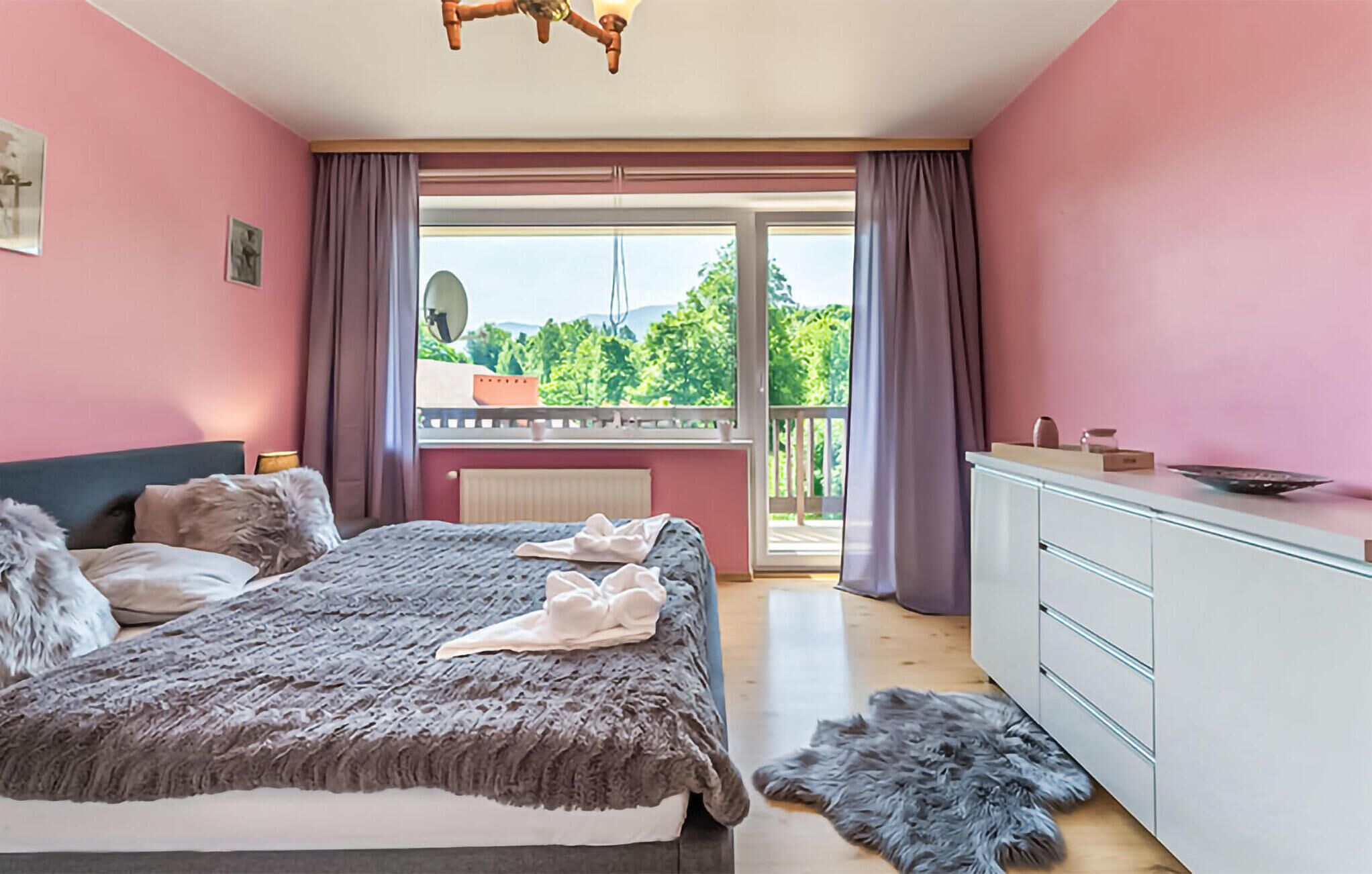 A bedroom with a bed, a dresser, and a balcony with a view of greenery.