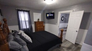 Een slaapkamer met een bed, een dressoir, een televisie aan de muur en een deur naar een andere kamer.