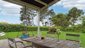 Ein Holztisch mit Stühlen unter einer überdachten Terrasse, versehen mit einer Hängeschaukel, umgeben von einem üppigen Grünbereich und hohen Bäumen.