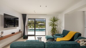 A modern living room with a teal sofa, a flat-screen TV, a large window overlooking a pool and greenery, and a potted plant. - Pet friendly home in Radosic (Radosic)