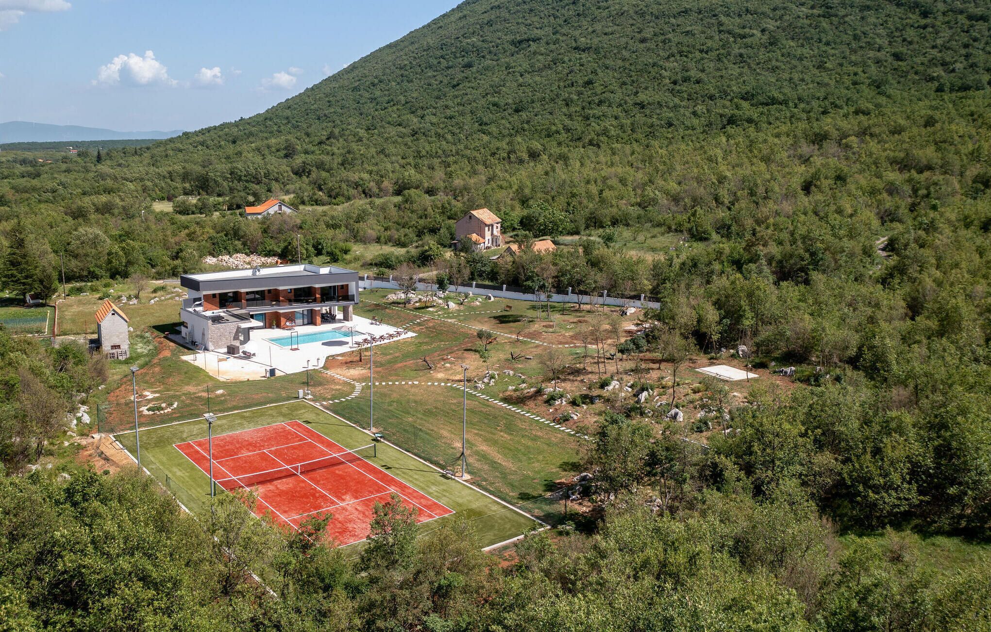 A modern house with a swimming pool, tennis court, and surrounding greenery.