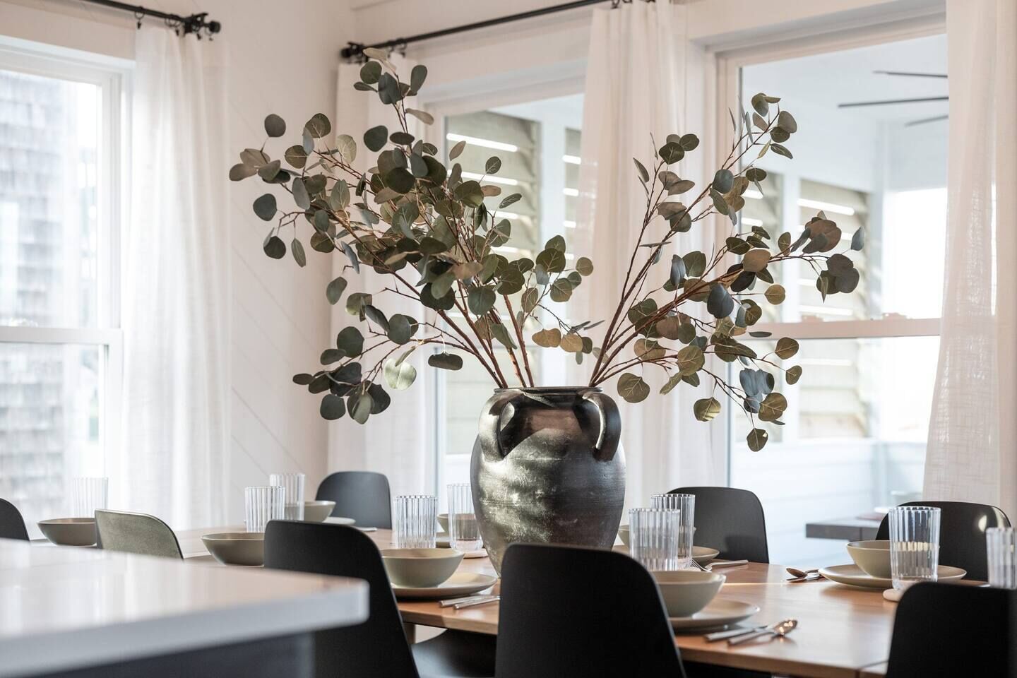 A dining area with a table set for two, featuring a vase with branches and leaves, and a cityscape view through large windows.